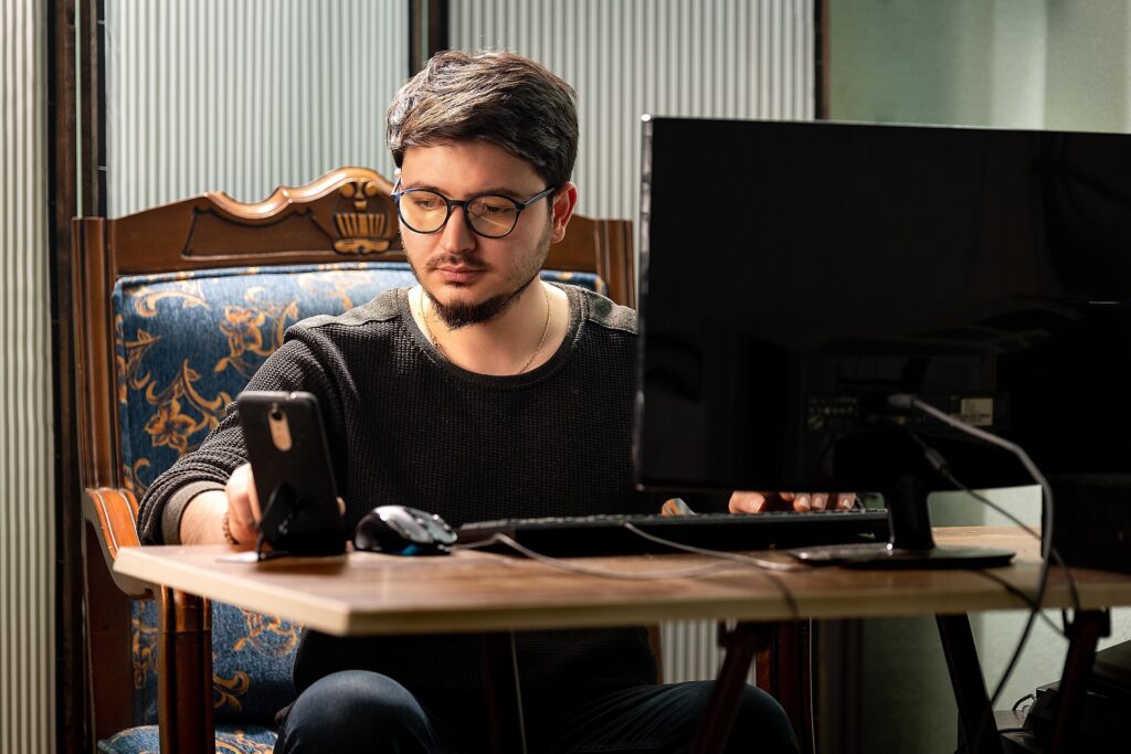 A man is sitting in front of his computer while using his mobile phone.